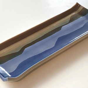 May include: A rectangular ceramic tray with curved, raised edges. The surface features a glossy glaze with horizontal bands of deep blue, muted brown, and light beige, creating a smooth, reflective finish across the elongated, shallow dish.
