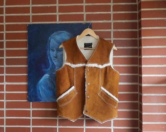 May include: A brown suede vest with a cream-colored shearling lining and trim. The vest has a V-neck, two front pockets, and is buttoned up. It hangs on a wooden hanger against a brick wall and a blue painting.