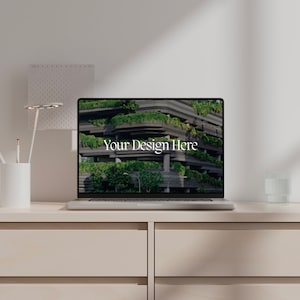 May include: A laptop computer with a black screen displaying the text "Your Design Here" against a background image of a building with green plants growing on the exterior. The laptop is sitting on a light brown wooden desk with a white pencil holder, a white lamp, and a clear glass on the surface.