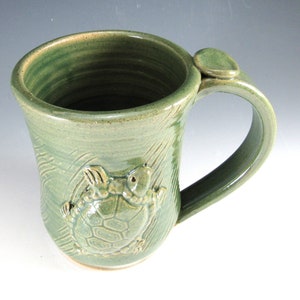 Puede incluir: Una taza de cerámica verde con un diseño de tortuga marina en relieve tridimensional. La taza tiene un asa y un patrón ondulado texturizado.
