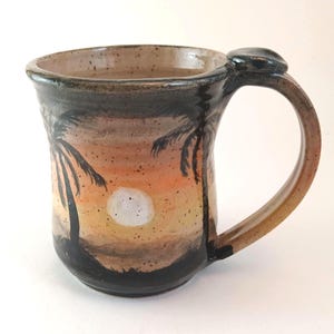 Puede incluir: Taza de cerámica pintada a mano con un diseño de puesta de sol y palmeras. La taza tiene un asa y borde marrón, con un acento negro en la parte superior. El diseño incluye tonos naranja, amarillo y negro, creando un ambiente tropical.