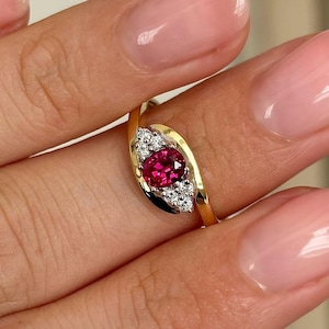 May include: A gold ring featuring a vibrant red gemstone, accented with smaller white gemstones. The ring's design is a flowing, asymmetrical shape, with the red stone as the focal point. The ring is set against a neutral background.