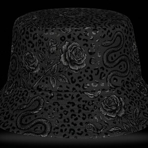 Puede incluir: Sombrero de pescador negro con un patrón repetido de rosas, serpientes y manchas de leopardo. El sombrero está hecho de una tela suave y tiene un ala ancha. El diseño es monocromático.