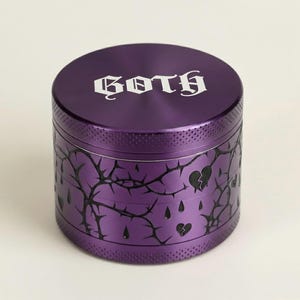 Puede incluir: Un molinillo cilíndrico morado con la palabra "GOTH" en letras góticas blancas en la tapa. El cuerpo presenta un diseño negro de espinas y corazones rotos. El molinillo es de metal.
