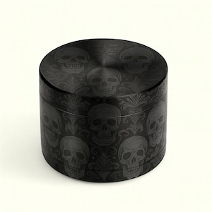 Peut inclure: Un broyeur cylindrique noir avec un motif de crâne et floral. Le dessus et les côtés du broyeur sont recouverts d'un motif répété de crânes et de motifs décoratifs. Le broyeur est fait d'un matériau sombre et mat.