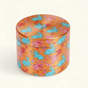 Könnte beinhalten: Ein runder, orangefarbener Metallbehälter mit einem sich wiederholenden Muster aus blauen Kaninchen, rosa Amoren, rosa Schlangen und Herzen. Der Behälter ist zylindrisch mit Deckel und Boden. Das Design ist skurril und farbenfroh.