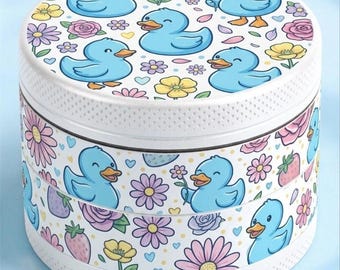 May include: A cylindrical metal herb grinder featuring a white base with a repeating pattern of light blue ducks, pink and yellow flowers, small hearts, and strawberries. The top lid and side sections display the same colorful graphic print.