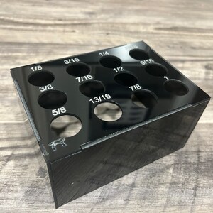 May include: Black acrylic drill bit organizer with 12 slots labeled with different sizes: 1/8, 3/8, 5/8, 3/16, 7/16, 13/16, 1/4, 1/2, 7/8, 9/16, 1, and 5/16.