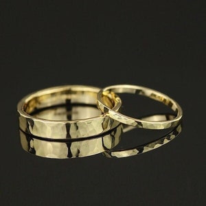 May include: Two gold hammered wedding bands, one wider than the other, are stacked on top of each other. The bands are on a black background.