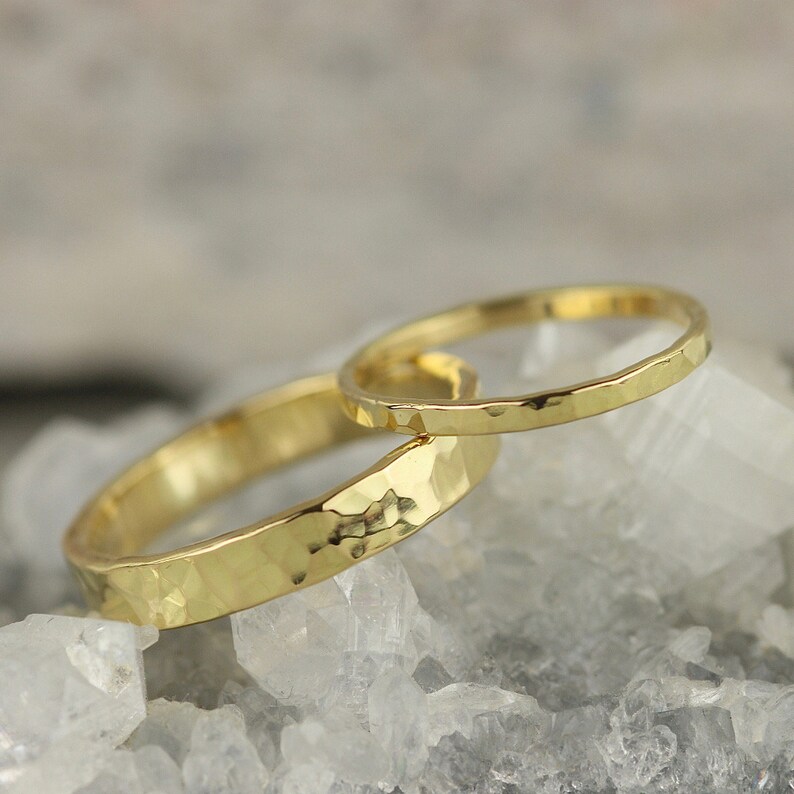 May include: Two gold hammered wedding bands, one wider than the other, resting on a bed of white crystals.