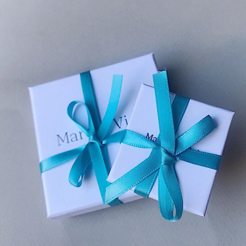 May include: Two white gift boxes tied with turquoise blue ribbon bows. The boxes are stacked on top of each other. The top box has the word "Mar" visible on the front. The bottom box has the word "Ma" visible on the front.