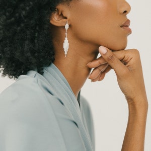 May include: A woman wearing a light blue shirt with silver geometric earrings. The earrings are long and dangle from her earlobes.