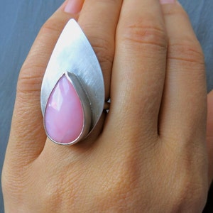 May include: A silver ring with a pink teardrop-shaped gemstone set in the center. The ring is designed with a leaf-like shape.