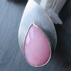 May include: A teardrop-shaped pink opal gemstone set in a silver pendant. The pendant has a smooth, brushed finish.