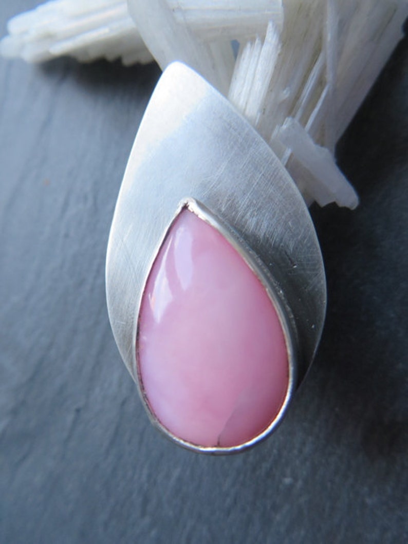 May include: A teardrop-shaped pink opal gemstone set in a silver pendant. The pendant has a smooth, brushed finish.