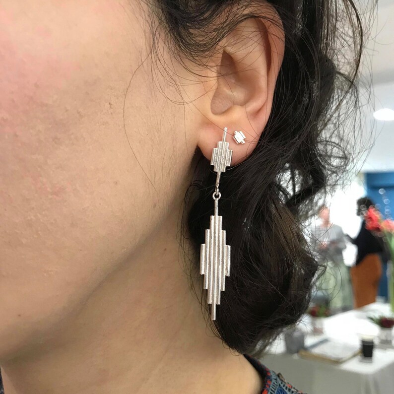 May include: Silver dangle earrings with a geometric design. The earrings are made of multiple thin, vertical bars that are stacked on top of each other to create a cascading effect. The earrings are a modern and stylish design.