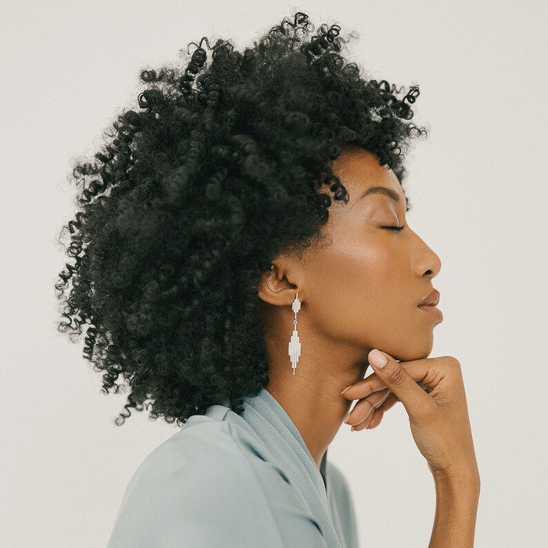 May include: A woman with curly black hair wearing a light blue shirt and silver dangle earrings. The earrings have a geometric design.