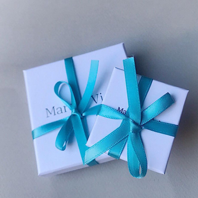 Puede incluir: Dos cajas de regalo blancas atadas con lazos de cinta azul turquesa. Las cajas est&aacute;n apiladas una encima de la otra. La caja superior tiene la palabra "Mar" visible en la parte delantera. La caja inferior tiene la palabra "Ma" visible en la parte delantera.