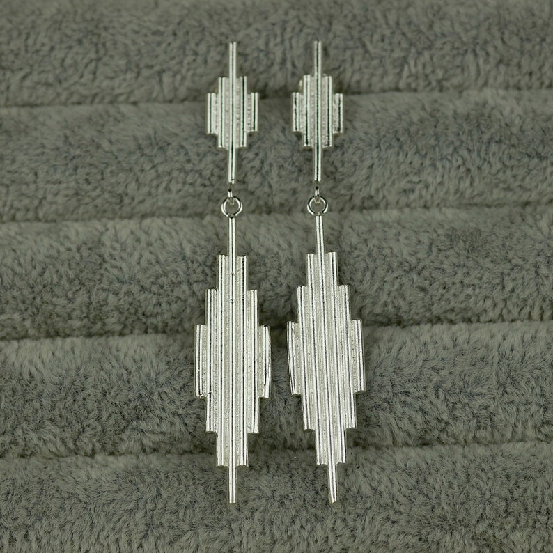 May include: A pair of silver geometric dangle earrings with a linear design. The earrings are long and narrow, with a pointed tip at the bottom.