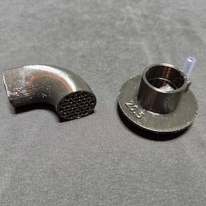 May include: Two black 3D-printed parts. One is a curved elbow with a honeycomb pattern on one end. The other is a cylindrical piece with a small tube attached, sitting on a circular base with the number "23.5" printed on it.