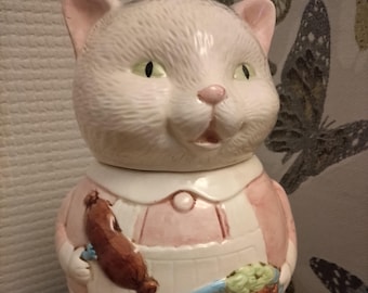 May include: A white ceramic cat figurine wearing a pink dress and holding a brown sausage. The cat has green eyes and a pink nose. The figurine is on a white pedestal with a price tag that reads "AD 301221 329".