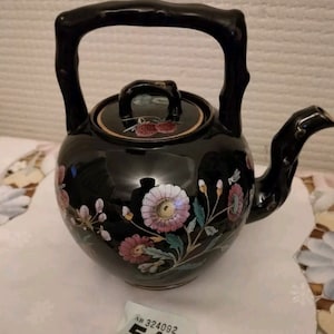 Puede incluir: Tetera negra con diseño floral y asa de bambú. La tetera tiene una tapa con ribete dorado. La tetera está sobre un mantel de encaje blanco. La etiqueta en el mantel dice "AB 324092 516".