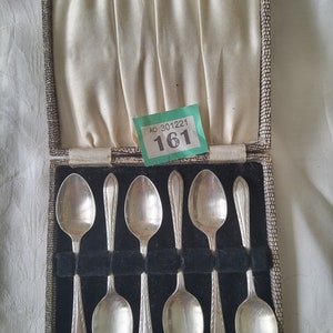 May include: A set of six silver spoons and two smaller silver pieces, displayed in a vintage case with a textured, patterned exterior. The spoons have a classic design, and the case has a cream-coloured satin lining.