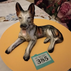 May include: A ceramic figurine of a German Shepherd dog lying down. The dog is painted in shades of brown, black, and white. The figurine is on a yellow circular base. A price tag with the number 385 is visible in the bottom right corner.