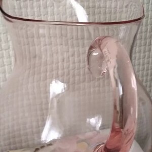 May include: A pink glass pitcher with a rounded body and a handle. The pitcher has a slightly frosted appearance.