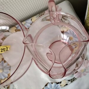 May include: A set of two pink glass bowls, one with a handle, both with a delicate, scalloped edge. The bowls are on a floral patterned surface.