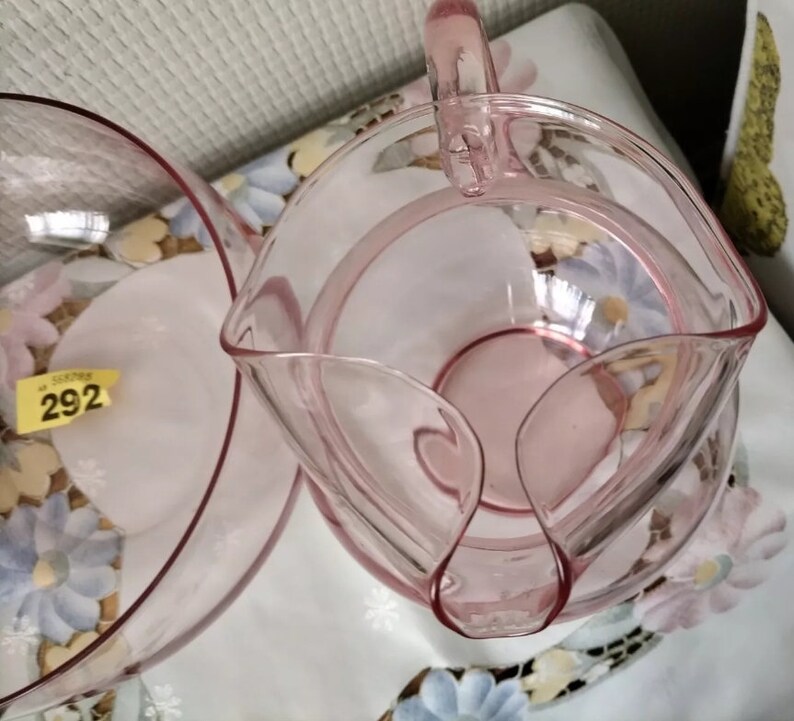 May include: A set of two pink glass bowls, one with a handle, both with a delicate, scalloped edge. The bowls are on a floral patterned surface.