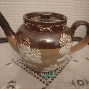 May include: A brown and cream ceramic teapot with a hunting scene design. The teapot has a lid with a knob on top and a spout on the side. The design features a man with a gun, a dog, and trees.