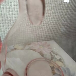 May include: A pink glass pitcher with a wide mouth and a handle. The pitcher is made of clear glass with a pink tint. The pitcher is sitting on a white surface with a floral pattern.
