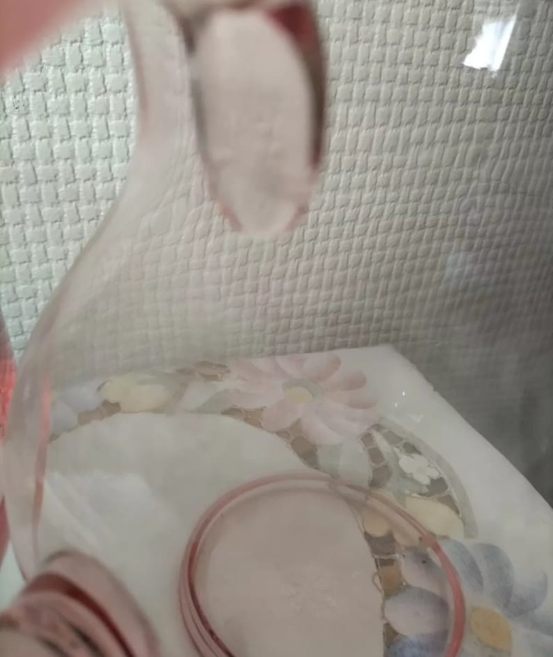 May include: A pink glass pitcher with a wide mouth and a handle. The pitcher is made of clear glass with a pink tint. The pitcher is sitting on a white surface with a floral pattern.
