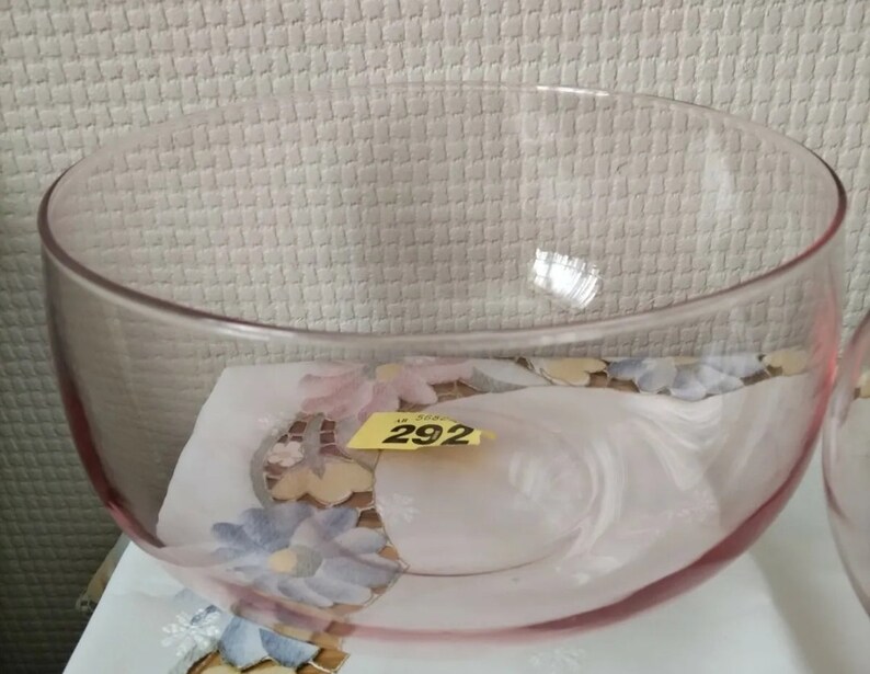 May include: A large, clear glass bowl with a pink tint. The bowl is sitting on a white surface with a floral pattern.