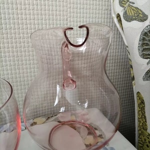 May include: A clear pink glass pitcher with a handle. The pitcher has a rounded body and a wide mouth.