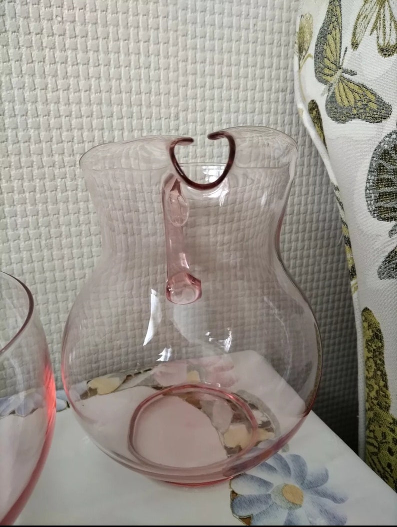 May include: A clear pink glass pitcher with a handle. The pitcher has a rounded body and a wide mouth.