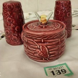 Op de afbeelding: Een set van drie keramische honingpotten met een bijendesign. De honingpot in het midden heeft een bij op de deksel. De honingpotten zijn versierd met een bloemenpatroon en zijn diep rood van kleur. Het prijskaartje geeft "AD 301221 139" aan.