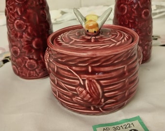 May include: A set of three ceramic honey pots with a bee design. The honey pot in the center has a bee on the lid. The honey pots are decorated with a floral pattern and are a deep red color. The price tag reads "AD 301221 139".