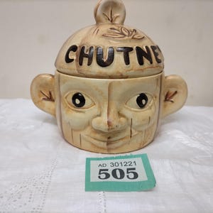 May include: A beige ceramic chutney jar with a lid, featuring a face design with large eyes, a nose, and a smiling mouth. The lid is inscribed with the word "CHUTNEY" in dark brown letters. The jar has ear-shaped handles.