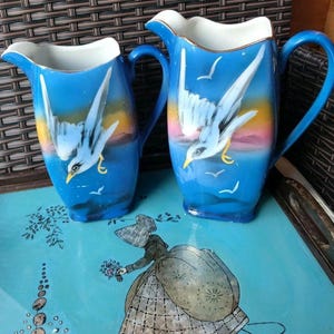 May include: Two blue ceramic jugs with white interiors and handles. Each jug features a painted design of a white bird in flight over a seascape with a sunset background. The jugs are on a blue tray with a woman illustration.