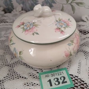 Puede incluir: Un tarro de cerámica blanco con un diseño floral y una pequeña figurita de ratón blanco en la tapa. El tarro tiene un borde verde alrededor de la parte superior e inferior. El tarro está sentado sobre un mantel de encaje blanco. La etiqueta en el tarro dice "AD 301221 132".