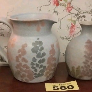 May include: Two ceramic vases with a floral design. The vases are a light blue colour with pink and green floral patterns. The larger vase has a handle and is shaped like a pitcher. The smaller vase is round. The vases are sitting on a wooden surface.