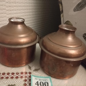 Peut inclure: Deux pots en cuivre antique avec couvercles. Les pots sont ronds et ont une lèvre légèrement évasée. Les couvercles sont bombés et ont un petit bouton sur le dessus. Les pots sont posés sur une table avec une nappe en dentelle blanche.