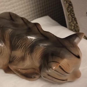 Op de afbeelding: Een keramiek beeldje van een slapende kat. De kat is bruin en zwart met een glanzende afwerking. Het beeldje ligt op zijn zij met zijn kop op zijn poten.
