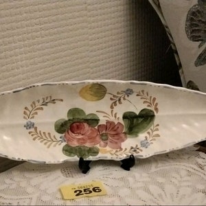 May include: White ceramic serving dish shaped like a leaf with a painted floral design. The dish has a raised edge and a scalloped rim. The flowers are pink, red, and yellow with green leaves. The dish is on a white lace tablecloth with a price tag that says "256".
