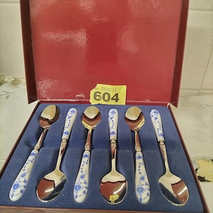 May include: A boxed set of six spoons. Each spoon features a silver bowl and a white handle adorned with blue floral designs. The spoons are presented in a red box with a blue velvet lining.