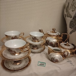 Puede incluir: Un juego de té vintage con detalles dorados, que incluye tazas, platillos, una tetera, una azucarera y una jarra de crema. El juego tiene un fondo blanco con un diseño floral. Las tazas están apiladas sobre los platillos.