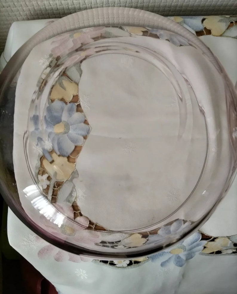 May include: A clear glass serving dish with a light pink rim. The dish is sitting on a white tablecloth with a floral pattern.