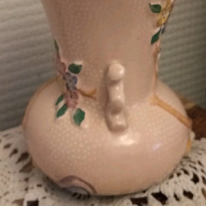 Może przedstawiać: Różowy ceramiczny wazon z kwiatowymi wzorami w odcieniach zieleni, żółci, błękitu i różu. Wazon ma fakturowaną powierzchnię i uchwyt po boku.